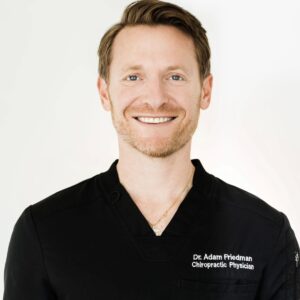 Close-up portrait of a smiling man in black medical scrubs with 'Dr. Adam Friedman Chiropractor Physician' embroidered on the chest.